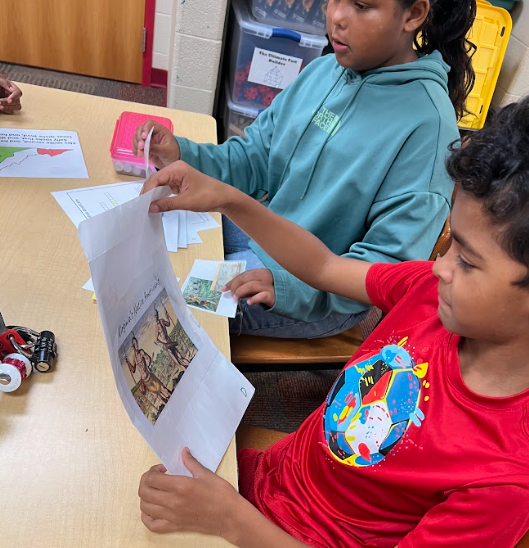 Two students sit at a table working with papers. One student in a red shirt holds up a sheet with an illustration and text while the other, wearing a green hoodie, looks on.