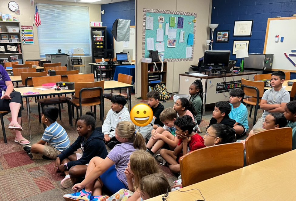 A group of elementary students sit cross-legged on the carpet in a classroom, attentively listening to a teacher who is seated in front of them. Behind the students are tables, chairs, and computer workstations.