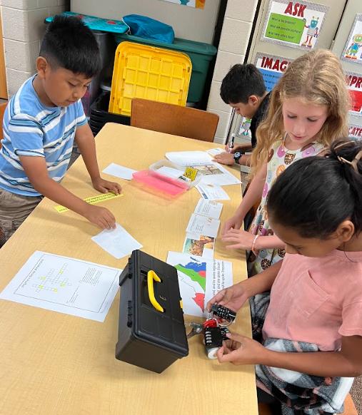 Four students stand around a table working on papers, maps, and a crossword puzzle. One student holds a lock on a black toolbox while others read clues.
