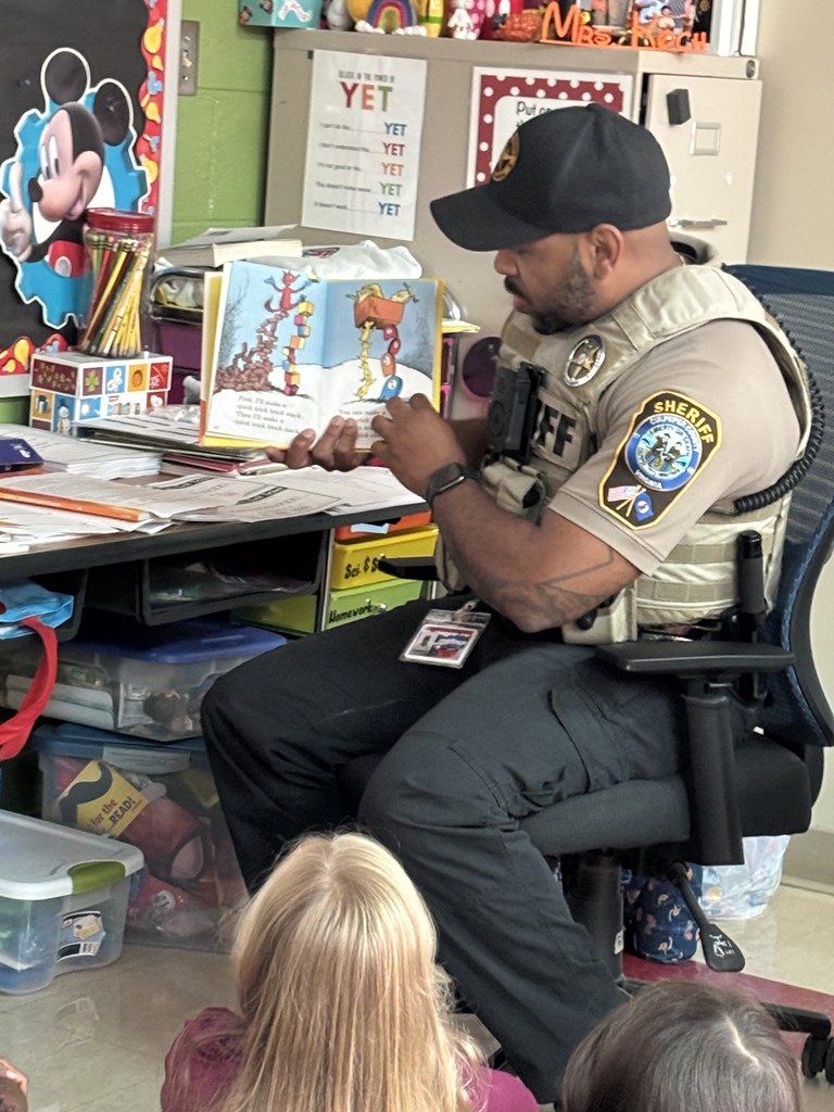 A close-up of the sheriff in uniform sitting at a classroom desk, holding open a colorful picture book and pointing to an illustration while reading to the children seated in front of him.