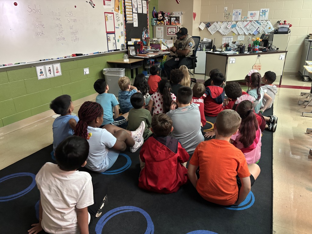 A group of young students sit cross-legged on a classroom rug, facing a uniformed sheriff who is seated at the teacher’s desk. The officer is reading aloud from a picture book while the children listen attentively.