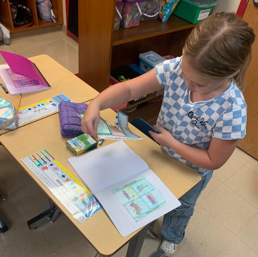 A student stands at her desk, holding crayons while working on a notebook activity that shows labeled sections for government branches.