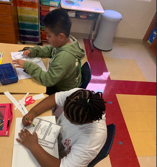 Two students sit side by side at a desk. One colors while the other cuts a foldable activity about the three branches of government.