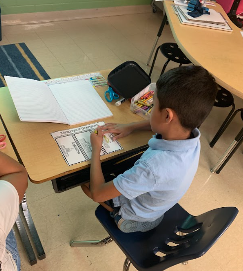 A boy colors a cut-out foldable titled “3 Branches of Government” at his desk with crayons and supplies nearby.