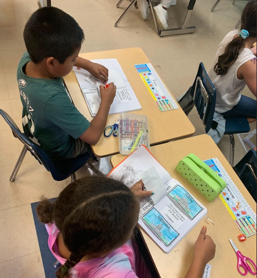 A boy and girl work at adjacent desks, coloring and gluing pieces of a foldable activity into their notebooks.