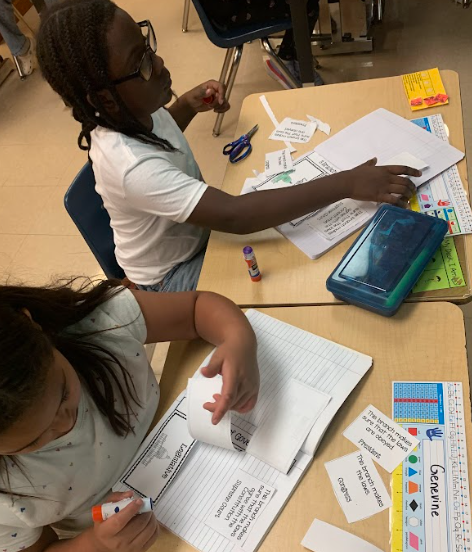 Two students glue and arrange cut-out pieces in their notebooks. Scissors, glue sticks, and activity sheets are on their desks.