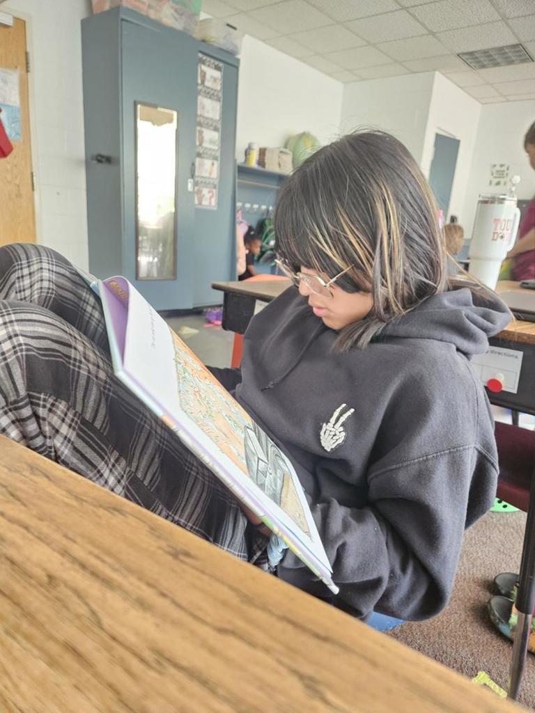 A student in glasses and a black hoodie sits on the floor near a desk, focused on reading a large picture book.