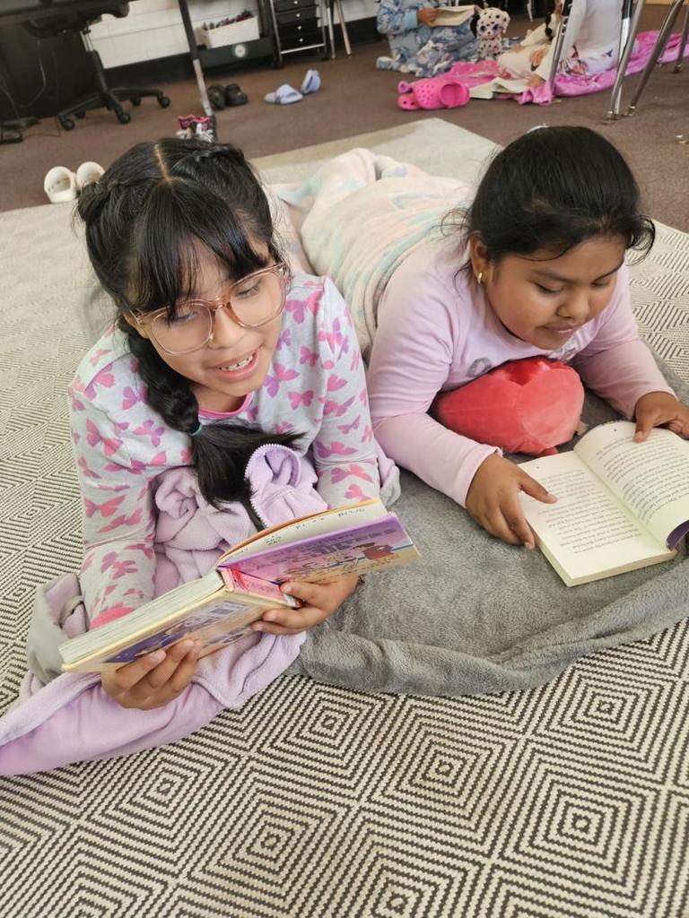 Two girls lying on a rug, smiling and reading chapter books. They are wrapped in blankets and look cozy in pajamas.