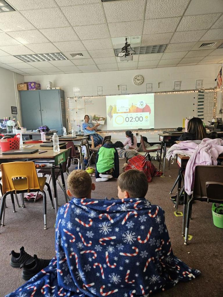 A classroom scene where a teacher is reading aloud from a book while students sit or lie on the floor with blankets. A large digital timer with “2:00” is projected on the board.