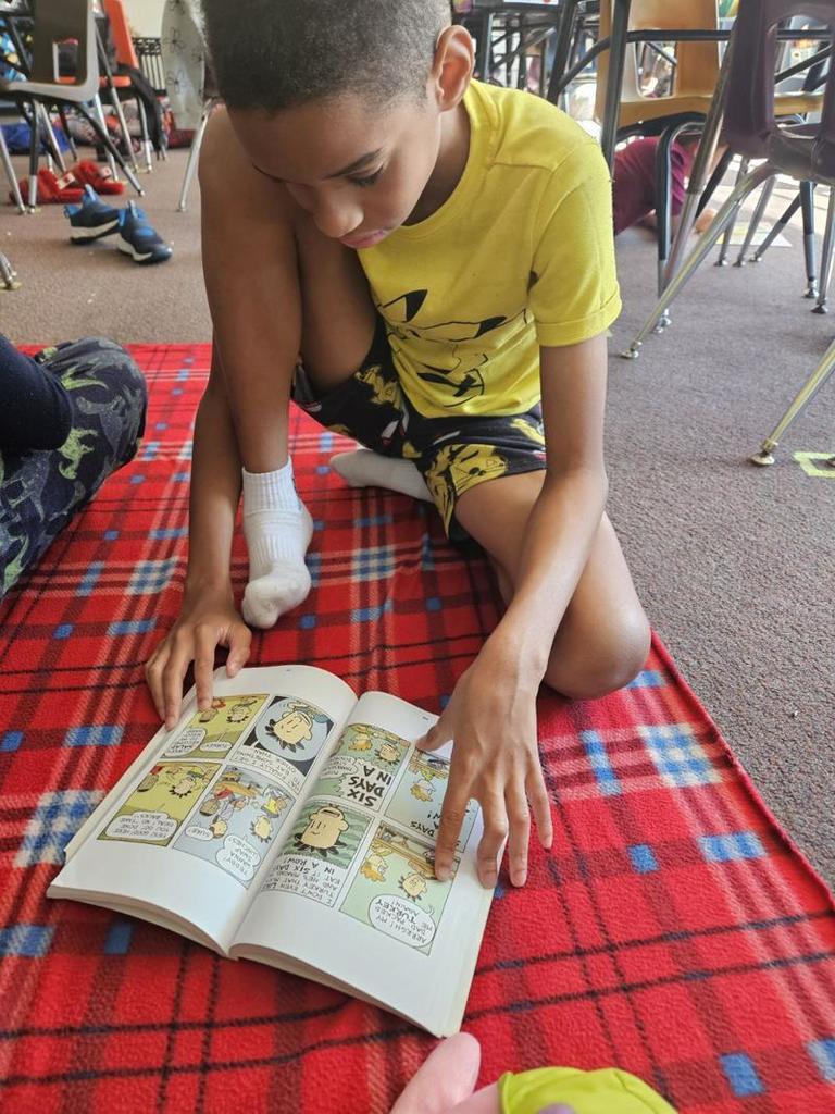 A boy in a yellow shirt and patterned shorts sits on a red plaid blanket, reading a comic-style book with colorful illustrations.