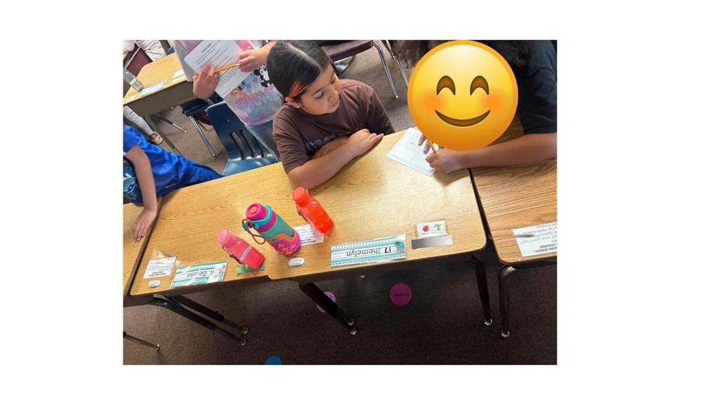 Two students sit at a desk; one writes on a paper while the other listens attentively, with water bottles lined up on the desk.