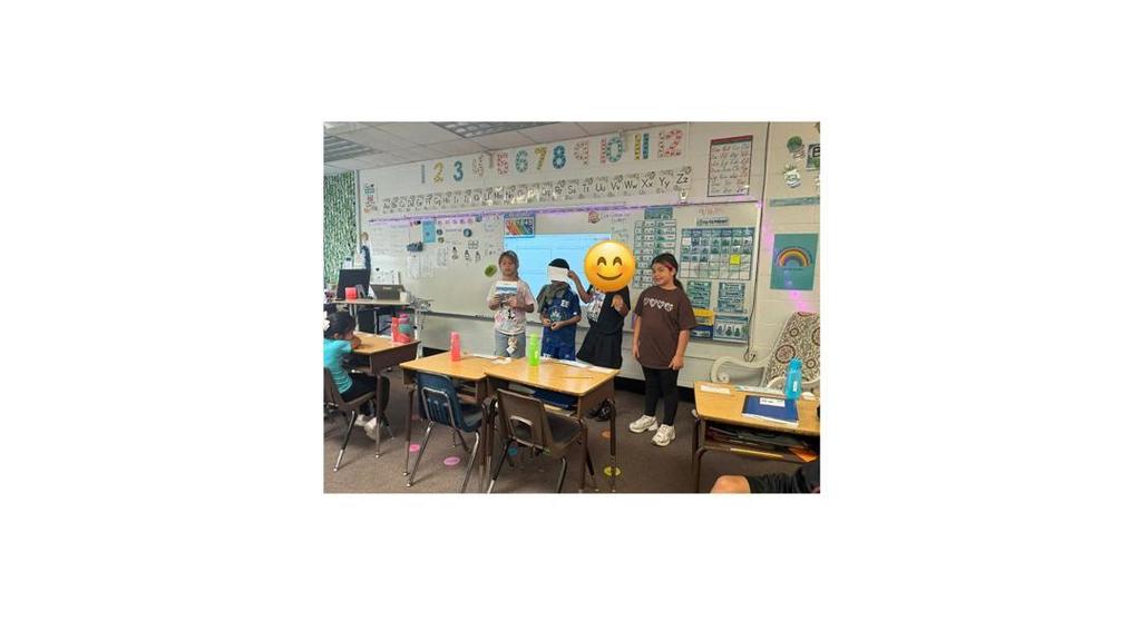 A group of four students stand at the front of the classroom presenting to their classmates, who sit at desks watching.