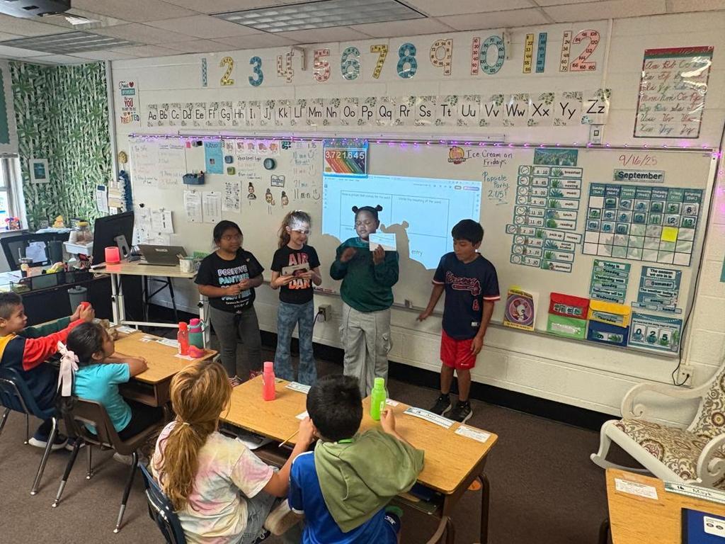 Four students present at the front of the classroom holding papers, while classmates at desks watch them.