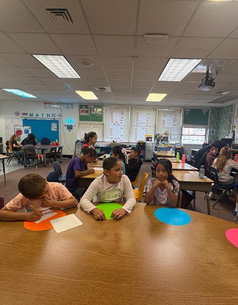 Three students sit at a round table with bright colored mats, while other students in the background work in groups.