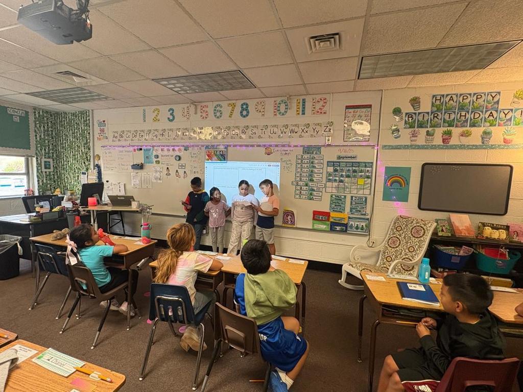 Another group of students presents at the front of the room near the whiteboard, while their classmates look on.