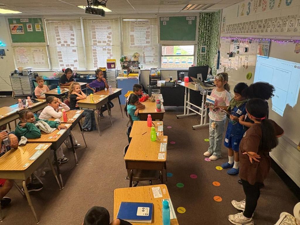 A group of students presents in front of the whiteboard, while the rest of the class listens from their desks.