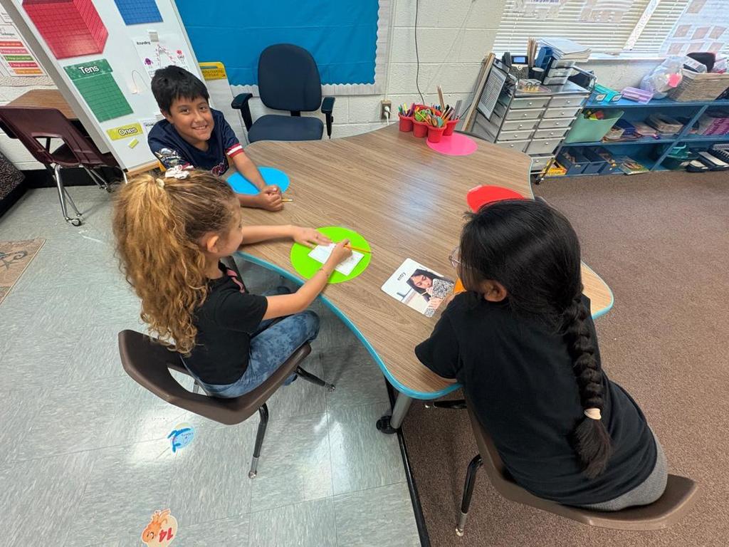 Three students sit at a smaller round table, discussing and writing on a paper, with colorful mats under their work.