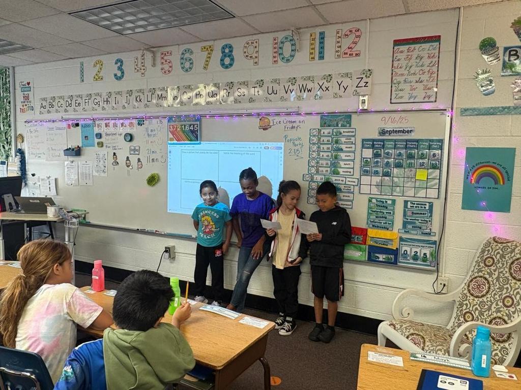 Four students present together at the front of the classroom, smiling as they share from their papers.