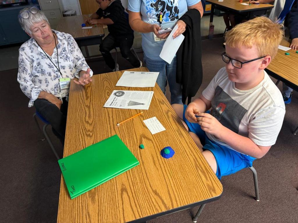 A student with glasses shapes blue and green modeling clay while an adult and another person observe nearby.
