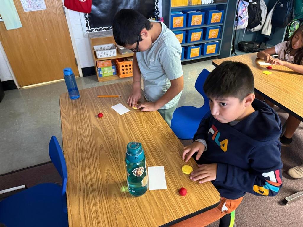 Two students at desks shape yellow and red modeling clay, with water bottles and pencils nearby.
