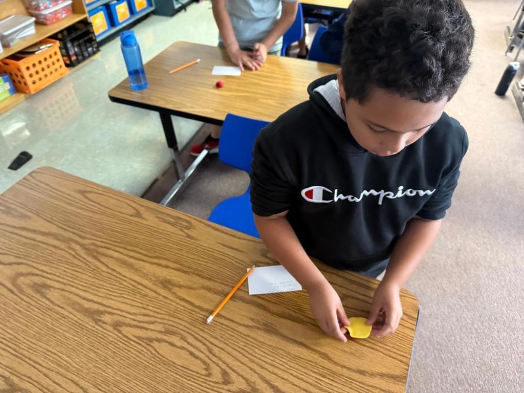 A student in a black hoodie works with yellow modeling clay at a desk with a pencil and paper.