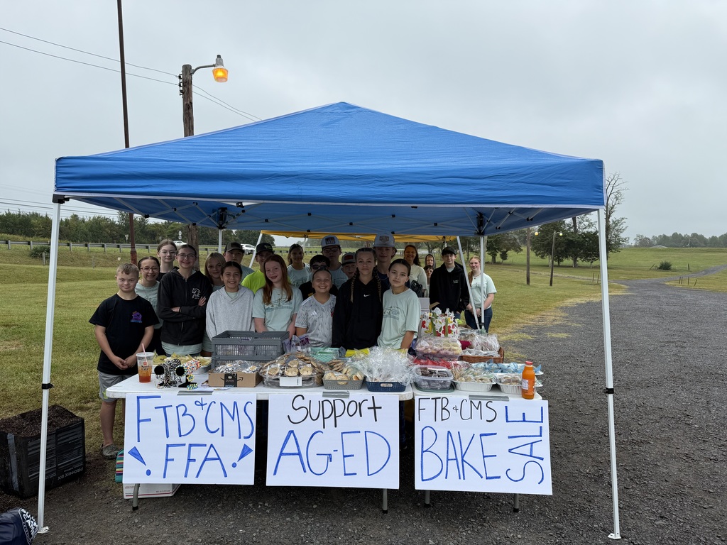 Alt text: A group of middle school FFA members stand under a blue canopy tent on a cloudy day, smiling behind tables filled with baked goods and drinks. Handwritten signs on the tables read “FTB+CMS FFA,” “Support AG-ED,” and “FTB+CMS Bake Sale.” The event is set up outdoors on a gravel path with grassy fields and trees in the background.