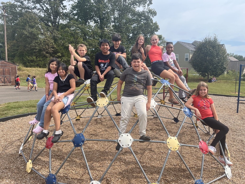 A group of ten 5th grade students smile and pose together on top of a metal dome climbing structure on the playground. Some sit near the top while others perch along the sides, enjoying recess.