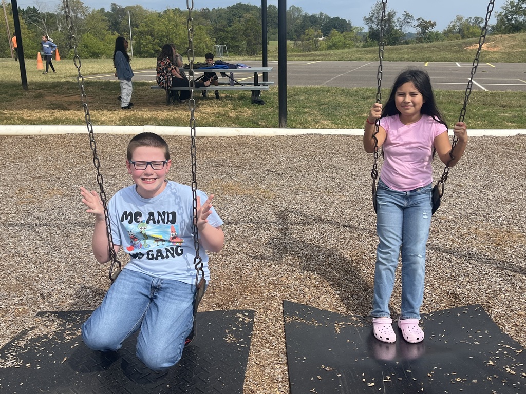 Two students are on swings. One boy in glasses and a SpongeBob shirt kneels on a swing smiling with hands raised, while a girl in a pink shirt and jeans sits on the swing next to him.