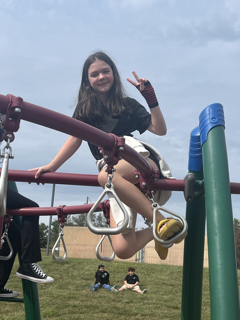 A girl in a black shirt and yellow sandals poses on the monkey bars, holding up a peace sign and smiling. Other students are visible sitting in the grass in the background.
