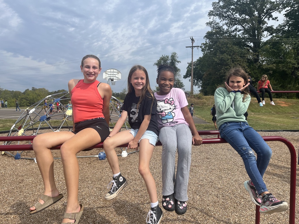 Four 5th grade girls sit together on a red bar structure at the playground. They are smiling and relaxed, with a basketball hoop and climbing dome visible in the background.