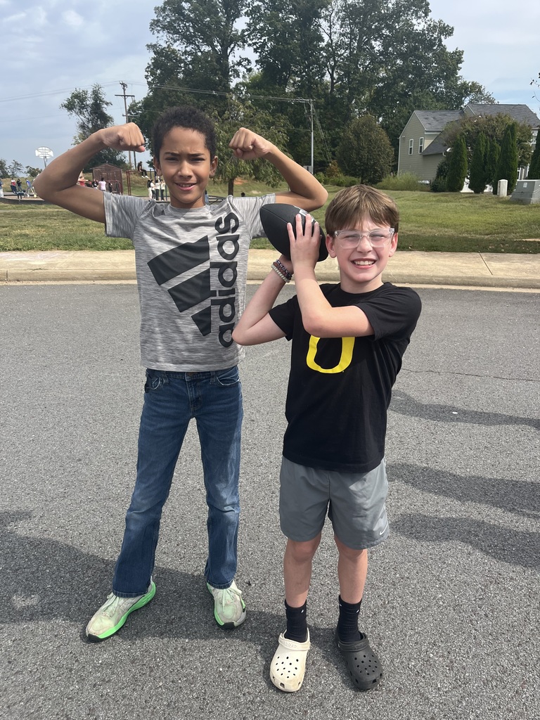 Two boys stand on the blacktop during recess. One flexes his arm muscles and smiles, wearing a gray Adidas shirt, while the other holds a football ready to throw and grins at the camera.