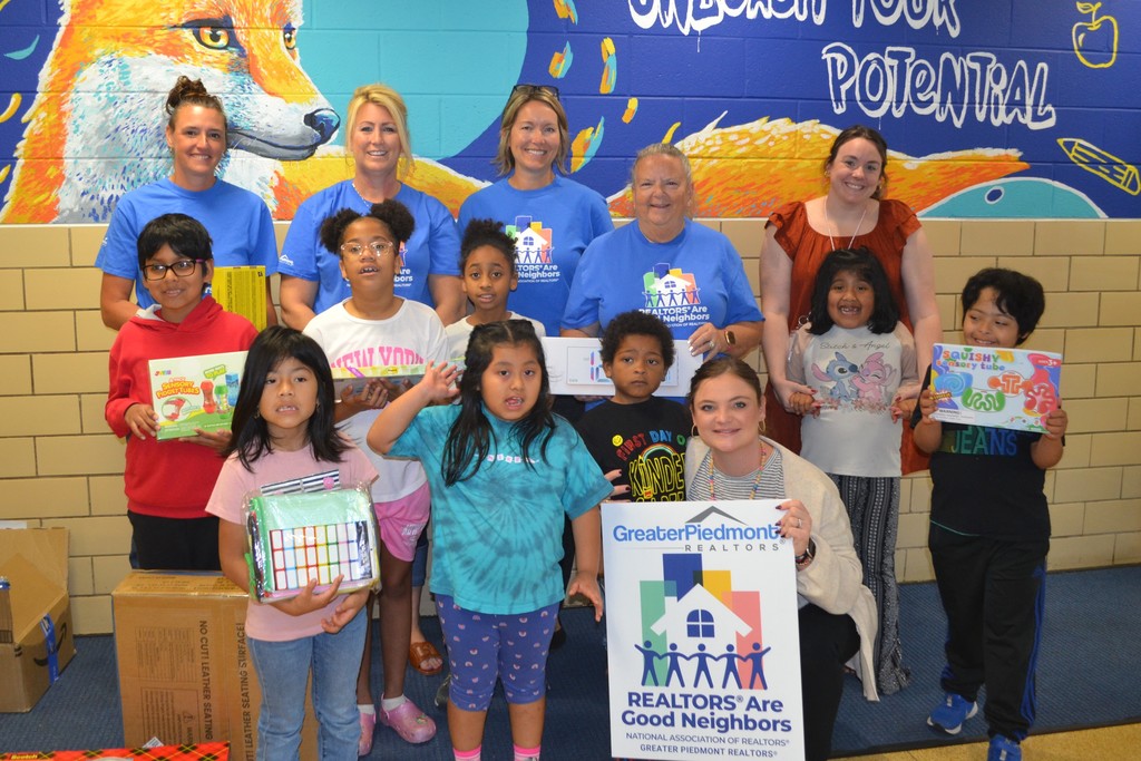 Image contains a group of students and their two teachers holding donated educational materials with 4 adults in matching blue tshirts.  Sign reads- Greater Piedmont Realtors Realtors are Good Neighbors