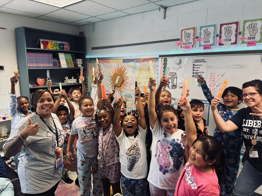A joyful group of elementary school students in a classroom, many wearing pajamas, smiling and holding colorful freeze pops in the air. Two teachers stand with them, also smiling and celebrating. The classroom has bright posters, bookshelves, and signs in both English and Spanish.