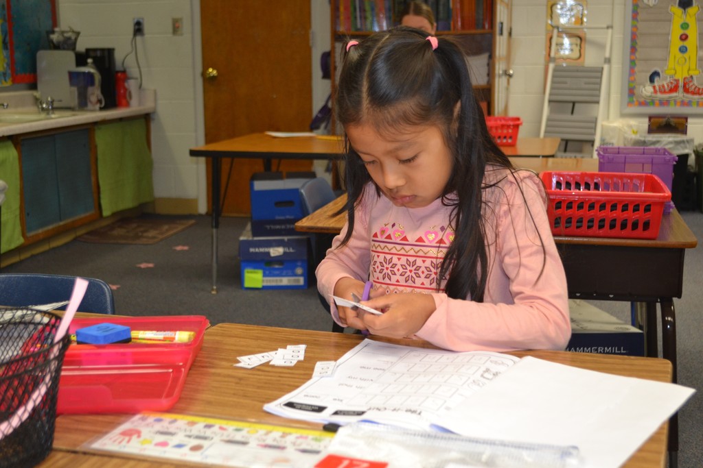 A student cutting little pieces of paper during a lesson while sitting at her desk in her school classroom.