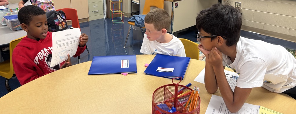 Three boys sit around a round table. One student in a red Spider-Man hoodie reads from a paper with a colorful design, while the other two lean in to listen. Blue folders and a red pencil caddy sit on the table.