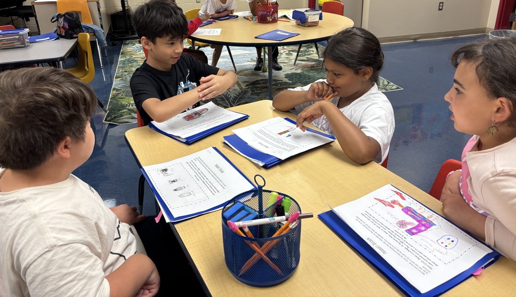 Four students sit at a round table, each with blue folders and papers. One student gestures with his hands while explaining his project, and the others listen closely. A blue supply caddy with markers and pencils is on the table.