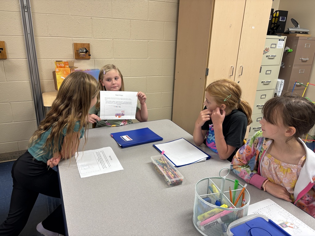 Four elementary students sit around a gray classroom table. A girl with blonde hair holds up a paper showing her project while three classmates watch. The table has blue folders, papers, and containers of crayons and markers.