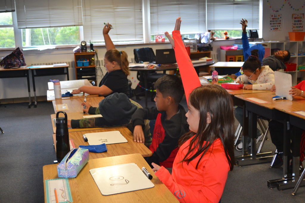 Image contains rows of desk with students, many students have their hands raised with math on their whiteboards.