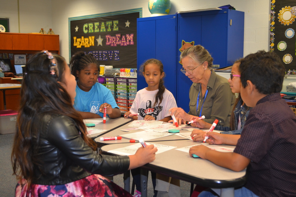 A small group of students are working at a table with the teacher.  Create Learn Dream Achieve is written on the back wall.  Students are using expo markers and whiteboards.