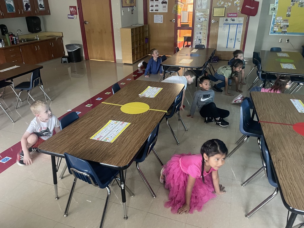 Children scattered across the classroom floor, balancing on their hands and feet in a crab-like position as part of a Cosmic Yoga exercise.