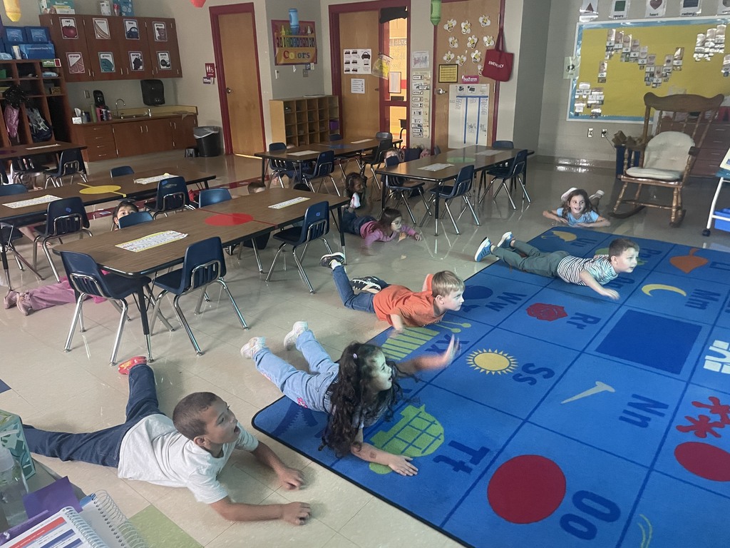 Several students lying on their stomachs on the classroom rug and floor, lifting their heads and arms as they follow a yoga pose from the Cosmic Yoga activity.