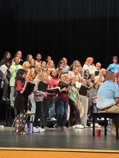 A group of middle and high school students stand on risers during a choir rehearsal on stage. Many hold black folders with sheet music while singing. They are dressed casually in colorful shirts and jeans, unlike a formal concert. In the foreground, part of an adult seated at a piano is visible, facing the students. The backdrop is a tall black curtain.