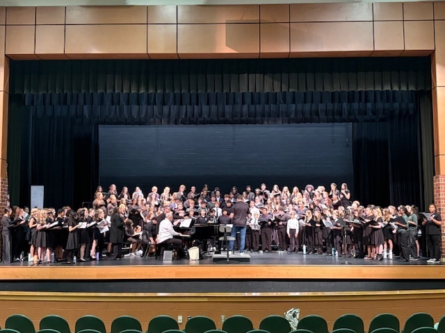 A large student choir rehearsal on a theater stage. The group, made up of dozens of students dressed in black concert attire, fills the entire width of the stage in several rows. A pianist sits at a grand piano on the left, while a conductor stands at the center facing the choir. The stage has a black backdrop, a high black curtain, and bright overhead lighting. In the foreground, empty green theater seats are visible.