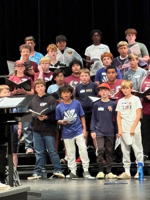 A group of middle school boys stand on risers singing in a choir rehearsal. Some hold music folders while others sing without them. They are dressed casually in t-shirts, jeans, and sneakers, with a piano and music stand visible at the front.
