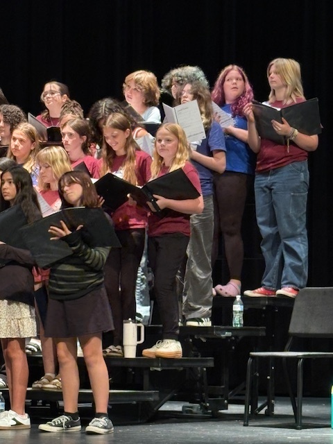 A close-up of a group of students standing on stage risers during choir rehearsal. They are holding black music folders while singing. Most are dressed casually in T-shirts, jeans, and sneakers, with some wearing matching maroon shirts. The black stage curtain serves as the backdrop.