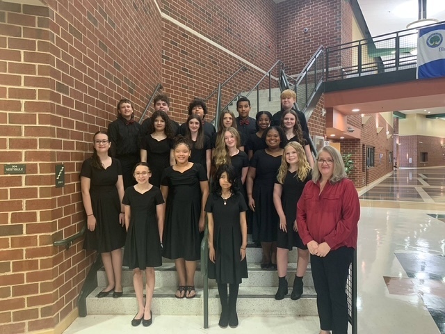 A group of students dressed in black performance attire stand together on a staircase inside a school building. They are posed in rows with their teacher, who is wearing a red blouse and black pants, standing in front to the right. The background shows brick walls, a stair railing, and a school hallway.