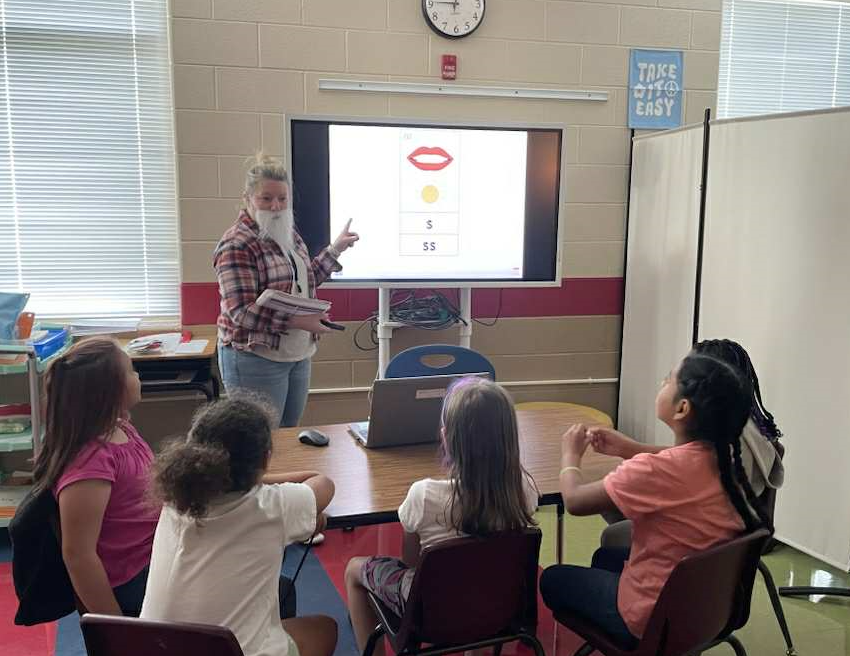 A teacher dressed in a plaid shirt and fake white beard stands at the front of a small group lesson, pointing to a screen with symbols while holding a book. Four elementary students sit at a round table watching and listening attentively.