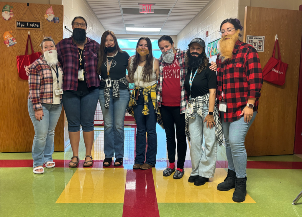A group of seven teachers stands together in a school hallway, smiling for the camera. They are dressed in flannel shirts, some with fake beards, celebrating Dress Like a Lumberjack Day.