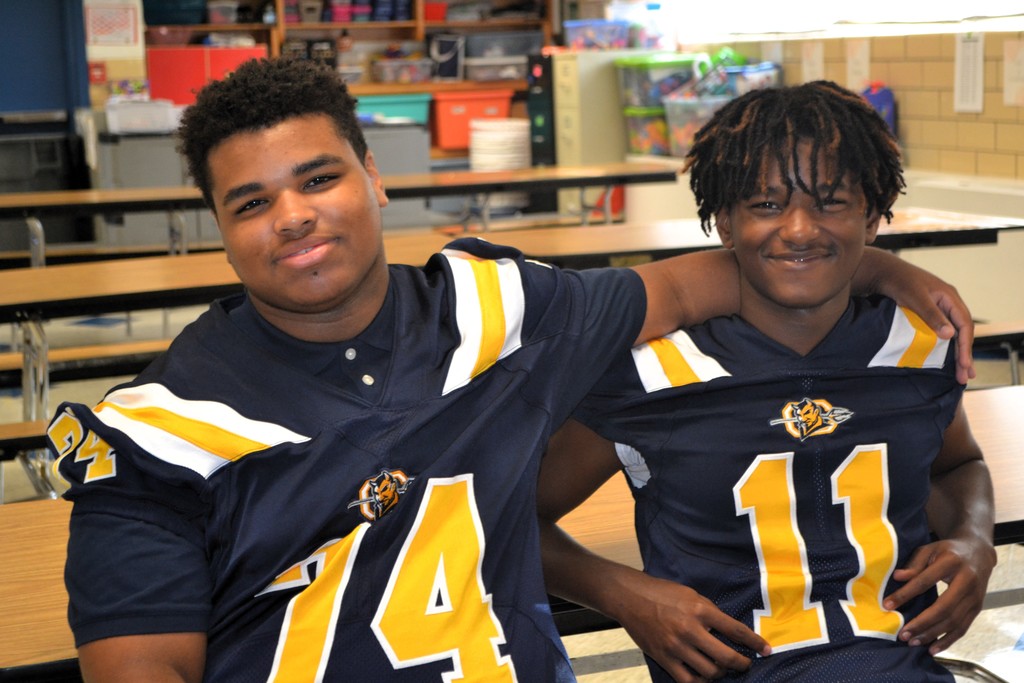 Two football players (74, 11) are smiling for a picture in the school cafeteria.