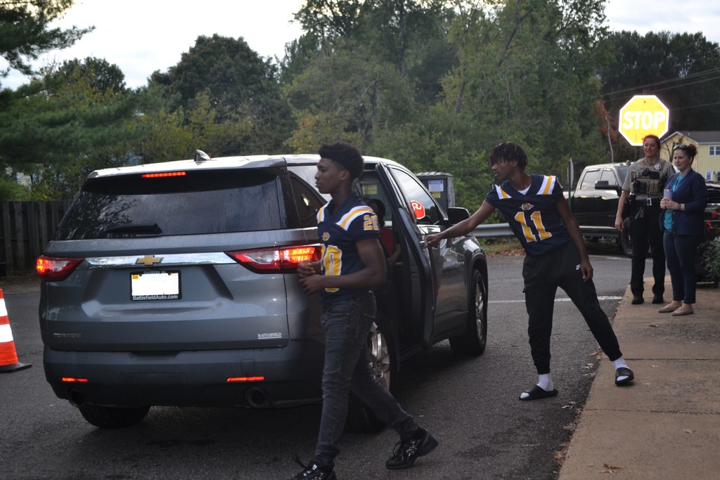 Two football players open the car door during school drop off to welcome the students.  Staff are in the background.